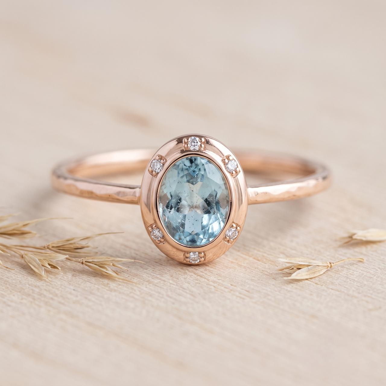 May include: A rose gold ring featuring an oval-cut light blue gemstone set in a bezel. The setting is accented with six small round white stones arranged around the central gem. The ring band has a textured, hammered finish throughout its surface.