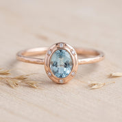 May include: A rose gold ring featuring an oval-cut light blue gemstone set in a bezel. The setting is accented with six small round white stones arranged around the central gem. The ring band has a textured, hammered finish throughout its surface.