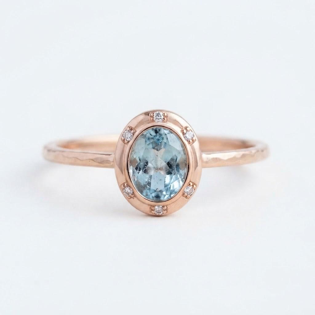 May include: A rose gold ring featuring a central oval-cut light blue gemstone surrounded by a halo of six small, clear round-cut stones. The band has a textured, hammered finish, providing a distinct contrast to the smooth, polished setting.
