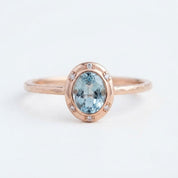 May include: A rose gold ring featuring a central oval-cut light blue gemstone surrounded by a halo of six small, clear round-cut stones. The band has a textured, hammered finish, providing a distinct contrast to the smooth, polished setting.