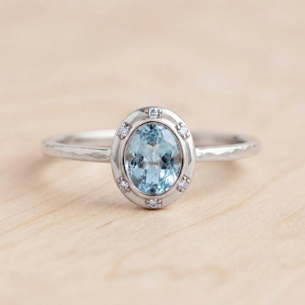 May include: A silver ring featuring a central oval-cut light blue gemstone surrounded by a halo of six small clear stones. The metal band has a textured, hammered finish, providing a distinct appearance to the silver setting and the ring structure.