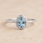 May include: A silver ring featuring a central oval-cut light blue gemstone surrounded by a halo of six small clear stones. The metal band has a textured, hammered finish, providing a distinct appearance to the silver setting and the ring structure.