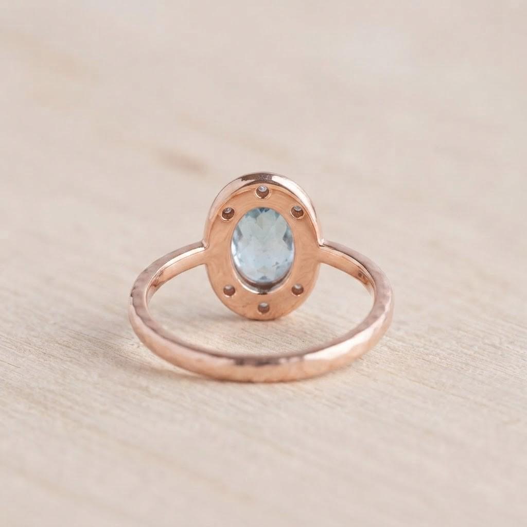 May include: A rose gold ring featuring an oval-shaped light blue gemstone set in a halo of small, clear stones. The metal band has a textured, hammered finish, and the setting is designed with a series of small circular cutouts around the center.