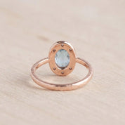 May include: A rose gold ring featuring an oval-shaped light blue gemstone set in a halo of small, clear stones. The metal band has a textured, hammered finish, and the setting is designed with a series of small circular cutouts around the center.