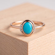 May include: A rose gold ring featuring a central oval-shaped turquoise cabochon stone. The setting includes three small round clear accent stones on each side of the central turquoise, integrated into the smooth, polished rose gold band design.