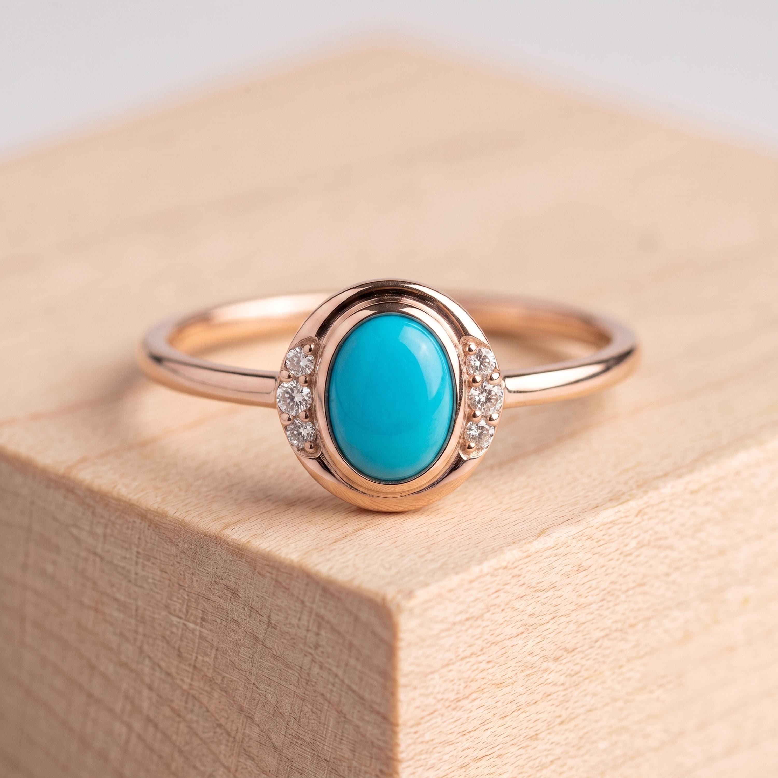 May include: A rose gold ring featuring a central oval-shaped turquoise cabochon stone. The setting includes three small round clear accent stones on each side of the central turquoise, integrated into the smooth, polished rose gold band design.
