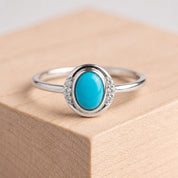 May include: A silver ring featuring a central oval-shaped turquoise gemstone in a smooth bezel setting. The ring band is accented on both sides of the center stone by three small, clear, round-cut white stones set into the silver metal frame.