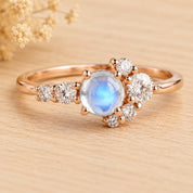 May include: A rose gold ring featuring a central round cabochon moonstone with a blue iridescent glow, flanked by a cluster of small, round, brilliant-cut white diamonds arranged in a delicate, asymmetrical floral-inspired pattern.