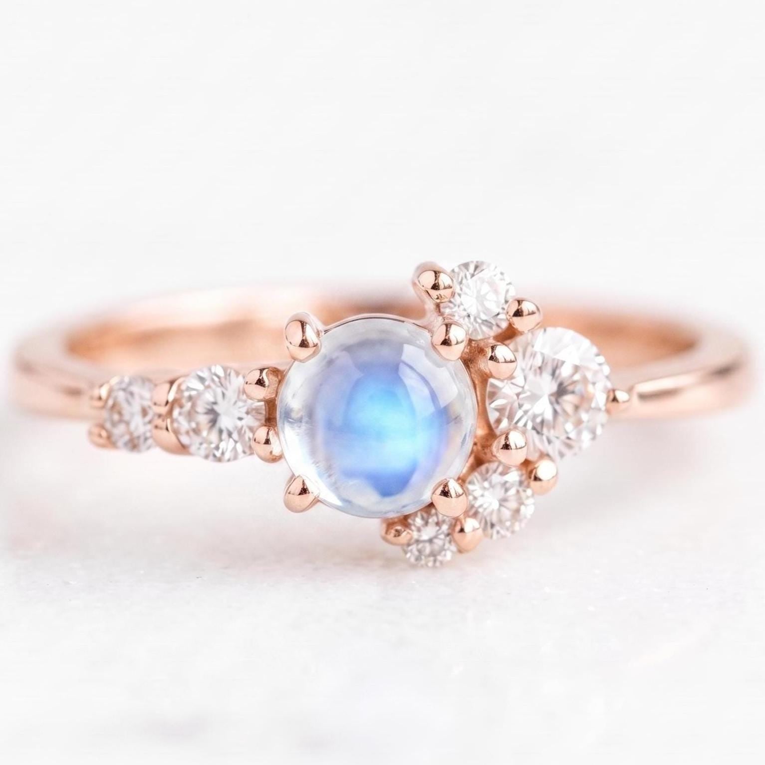 May include: A rose gold ring featuring a central round cabochon moonstone with a blue sheen. The stone is flanked by a cluster of small, round, clear faceted diamonds arranged in a graduated pattern along the band on both sides.