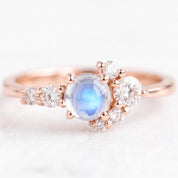 May include: A rose gold ring featuring a central round cabochon moonstone with a blue sheen. The stone is flanked by a cluster of small, round, clear faceted diamonds arranged in a graduated pattern along the band on both sides.