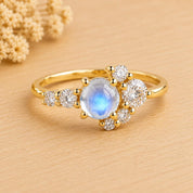 May include: A gold ring featuring a central round cabochon moonstone with a blue iridescent glow, surrounded by a cluster of small, clear, round-cut white stones arranged in an asymmetrical pattern along the polished gold band.