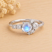 May include: A silver-toned ring featuring a central round cabochon moonstone with a blue iridescent glow. The stone is surrounded by a cluster of seven small, clear, round-cut faceted stones arranged in a asymmetrical pattern along the band.