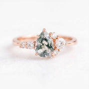May include: A rose gold ring featuring a central pear-shaped moss agate stone with dark green dendritic inclusions. The stone is flanked by a cluster of small, round, clear white diamonds set along the band on both sides of the main setting.