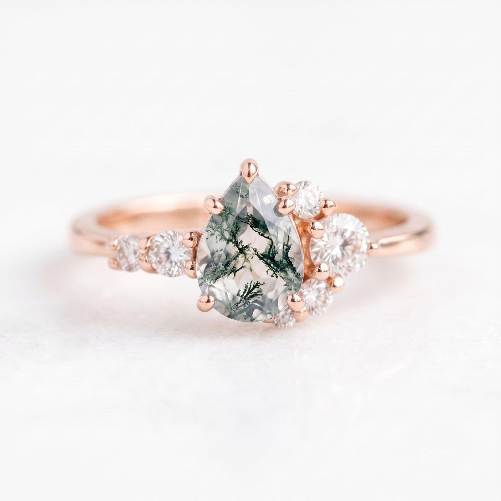 May include: A rose gold ring featuring a central pear-shaped moss agate stone with dark green dendritic inclusions. The stone is flanked by a cluster of small, round, clear white diamonds set along the band on both sides of the main setting.