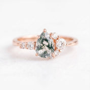 May include: A rose gold ring featuring a central pear-shaped moss agate stone with dark green dendritic inclusions. The stone is flanked by a cluster of small, round, clear white diamonds set along the band on both sides of the main setting.