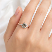 May include: A rose gold ring featuring a central pear-shaped moss agate stone surrounded by a cluster of small, clear round-cut accent stones on a thin, polished metal band. The design highlights the green inclusions within the gemstone.