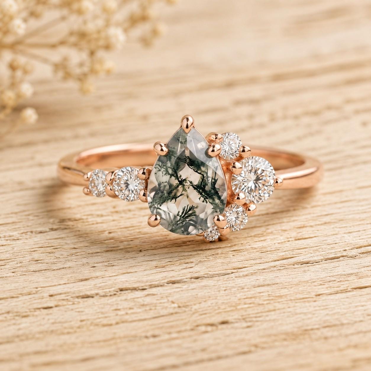 May include: A rose gold ring featuring a pear-shaped moss agate center stone with dark green dendritic inclusions. The setting is accented by a cluster of small, round, clear white diamonds arranged along the band and around the central stone.