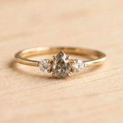 May include: A gold ring featuring a central pear-shaped salt and pepper diamond flanked by two smaller round brilliant-cut diamonds. The stones are set in a prong setting on a thin, polished gold band, creating a delicate three-stone design.