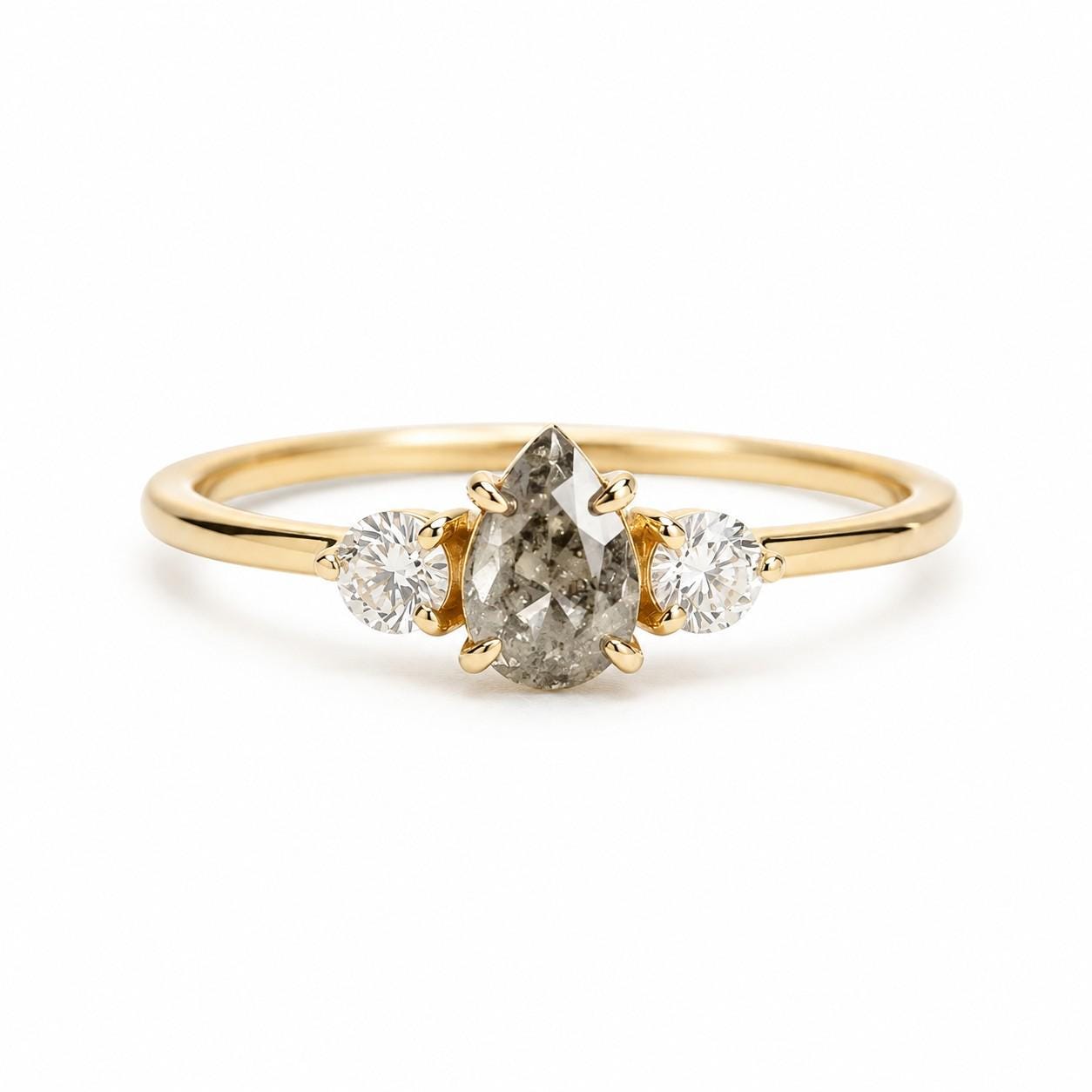 May include: A gold ring featuring a central pear-shaped salt and pepper diamond held by prongs, flanked by two smaller round brilliant-cut white diamonds. The band is smooth and polished, showcasing a classic three-stone design with a yellow gold setting.