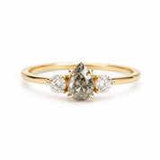 May include: A gold ring featuring a central pear-shaped salt and pepper diamond held by prongs, flanked by two smaller round brilliant-cut white diamonds. The band is smooth and polished, showcasing a classic three-stone design with a yellow gold setting.