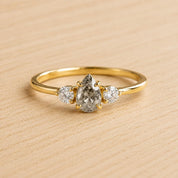 May include: A gold ring featuring a central pear-shaped salt and pepper diamond flanked by two smaller round brilliant-cut white diamonds. The stones are set in a yellow gold band with a polished finish, showcasing a classic three-stone design.