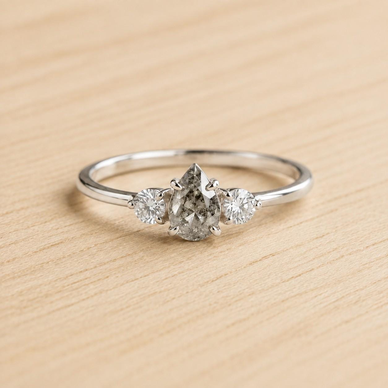 May include: A silver-toned ring featuring a central pear-shaped salt and pepper diamond flanked by two smaller round brilliant-cut white diamonds. The stones are set in a prong setting on a thin, polished metal band with a smooth, reflective finish.