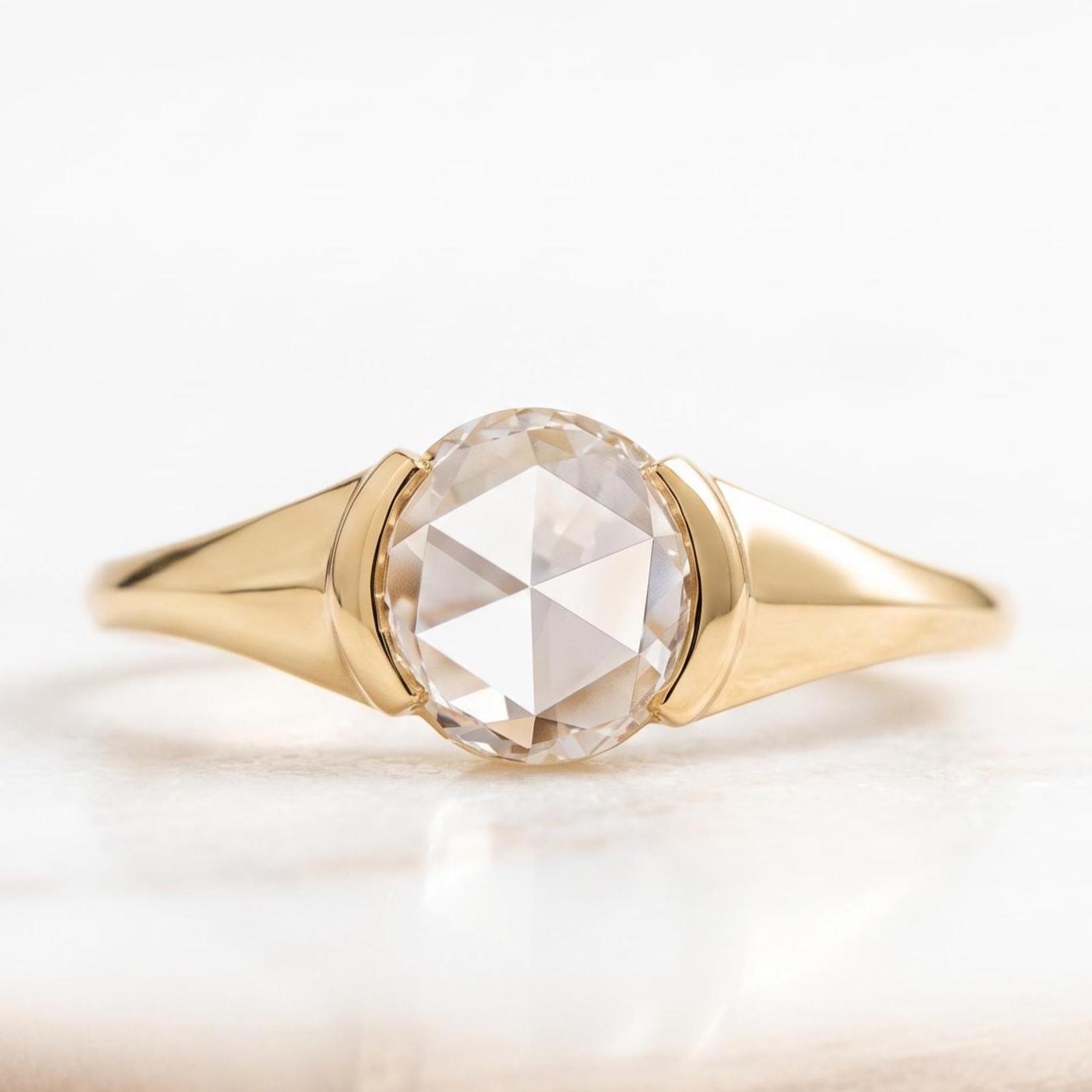 May include: A gold ring featuring a central round rose-cut gemstone with a faceted surface. The band is crafted from polished yellow gold, tapering smoothly toward the sides, creating a sleek and continuous metal frame around the stone.