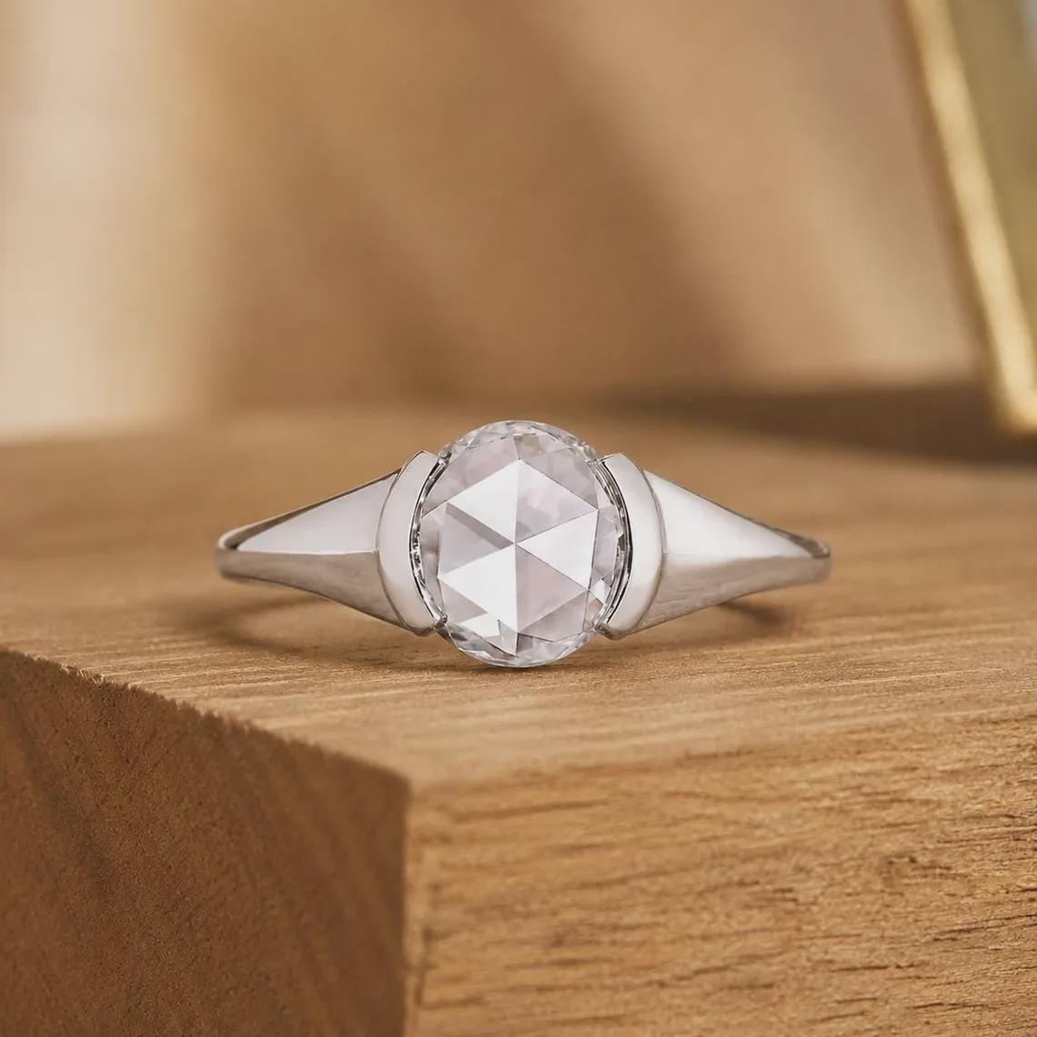 May include: A silver-toned ring featuring a central, round, rose-cut clear gemstone. The stone is held in a bezel setting that transitions into smooth, tapered metal shoulders. The band has a polished finish and a clean, symmetrical design.