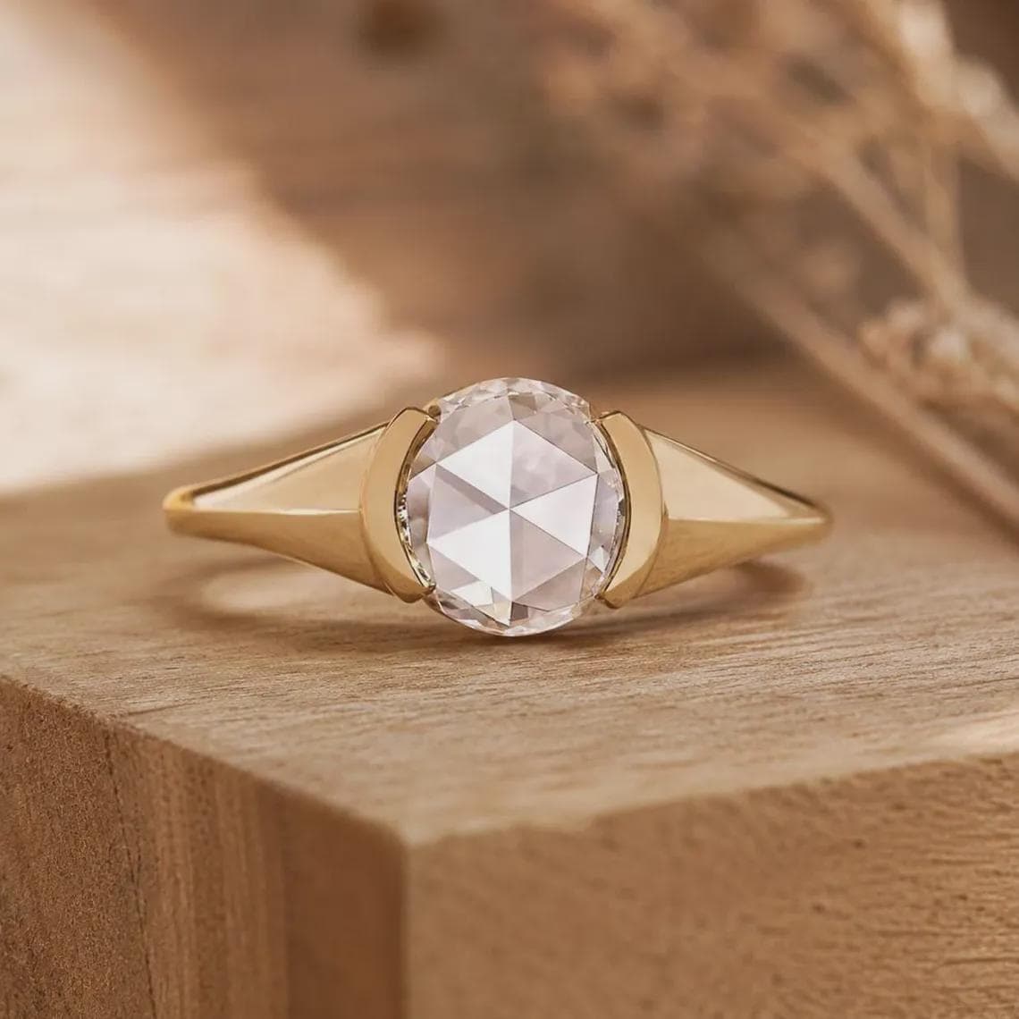 May include: A gold ring featuring a central round rose-cut clear gemstone held in a bezel setting. The band has a smooth, polished finish that tapers toward the sides, creating a clean, geometric look with a bright, reflective metallic surface.