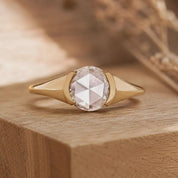 May include: A gold ring featuring a central round rose-cut clear gemstone held in a bezel setting. The band has a smooth, polished finish that tapers toward the sides, creating a clean, geometric look with a bright, reflective metallic surface.