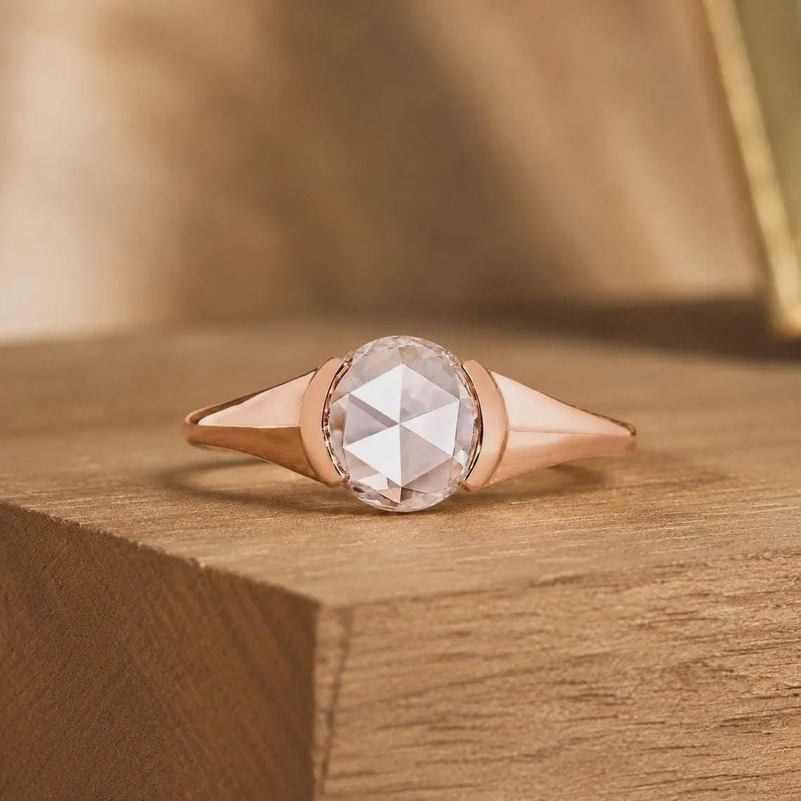 May include: A rose gold ring featuring a central round rose-cut gemstone with a faceted surface. The band has a smooth, polished finish that tapers toward the sides, creating a sleek and continuous metal frame around the clear, translucent stone.