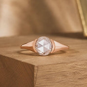 May include: A rose gold ring featuring a central round rose-cut gemstone with a faceted surface. The band has a smooth, polished finish that tapers toward the sides, creating a sleek and continuous metal frame around the clear, translucent stone.