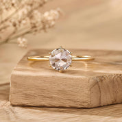 A diamond engagement ring with a large, clear gemstone is displayed on a wooden surface, with a blurred background.