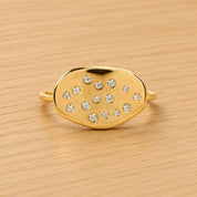 A gold ring with a large oval-shaped stone in the center, surrounded by smaller stones, sits on a wooden surface.