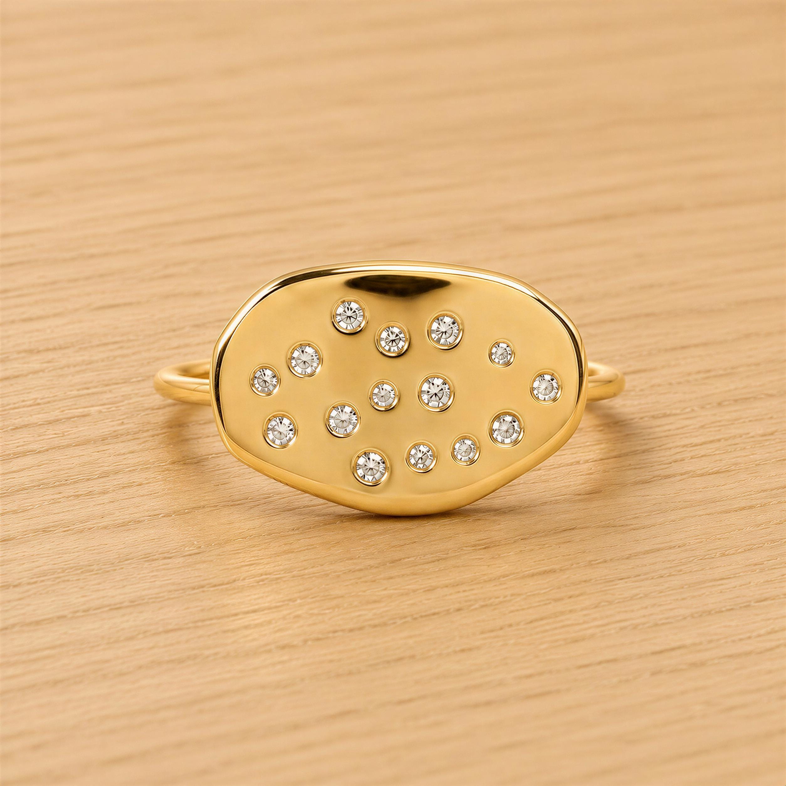 A gold ring with a large oval-shaped stone in the center, surrounded by smaller stones, sits on a wooden surface.