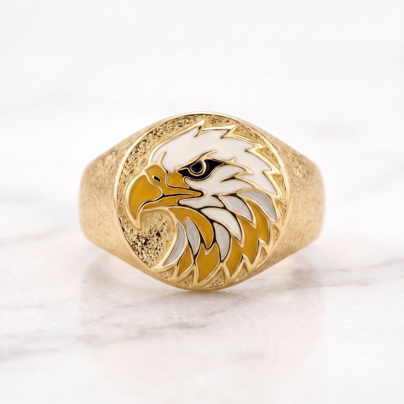 May include: A gold-toned ring featuring a circular face with a textured background. The center displays an enamel inlay of an eagle head in profile, rendered in white, yellow, and black colors. The band has a matte, granular surface texture.