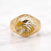 May include: A gold-toned ring featuring a circular face with a textured background. The center displays an enamel inlay of an eagle head in profile, rendered in white, yellow, and black colors. The band has a matte, granular surface texture.