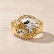 May include: A gold-toned ring featuring a circular face with an inlaid enamel design of an eagle head. The eagle is depicted in profile with white feathers, a yellow beak, and a black eye. The band has a textured, matte finish throughout.
