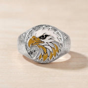 May include: A silver-toned ring featuring a circular face with an inlaid eagle head design. The eagle is rendered in white and yellow enamel with a black eye detail. The surrounding metal surface has a textured, matte, and speckled finish.