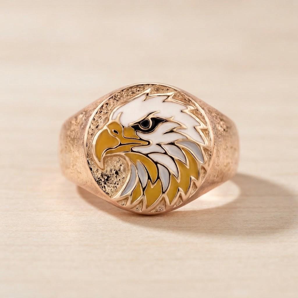 May include: A rose gold-toned ring featuring a circular face with an inlaid enamel design of an eagle head. The eagle is depicted in profile with white feathers, a yellow beak, and a black eye. The band has a textured, matte finish throughout.