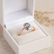 May include: A rose gold ring featuring an oval-cut light blue gemstone set in a bezel setting, surrounded by a halo of small clear stones on a thin band, presented inside a white jewelry box with a soft cream-colored velvet interior lining.