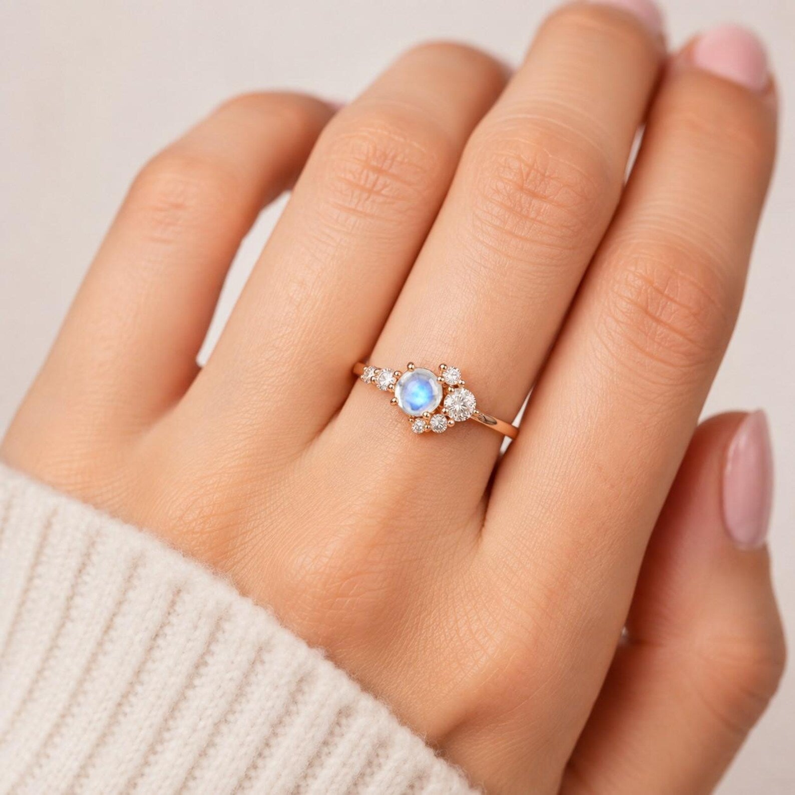 May include: A rose gold ring featuring a central round iridescent moonstone flanked by a cluster of smaller clear round stones on each side. The band is thin and metallic, showcasing a delicate arrangement of gemstones set in a prong style.