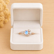 May include: A rose gold ring featuring a central round moonstone cabochon surrounded by a cluster of small, clear, round-cut accent stones. The ring is displayed inside an open, white, rectangular jewelry box with a soft, velvet-like interior.