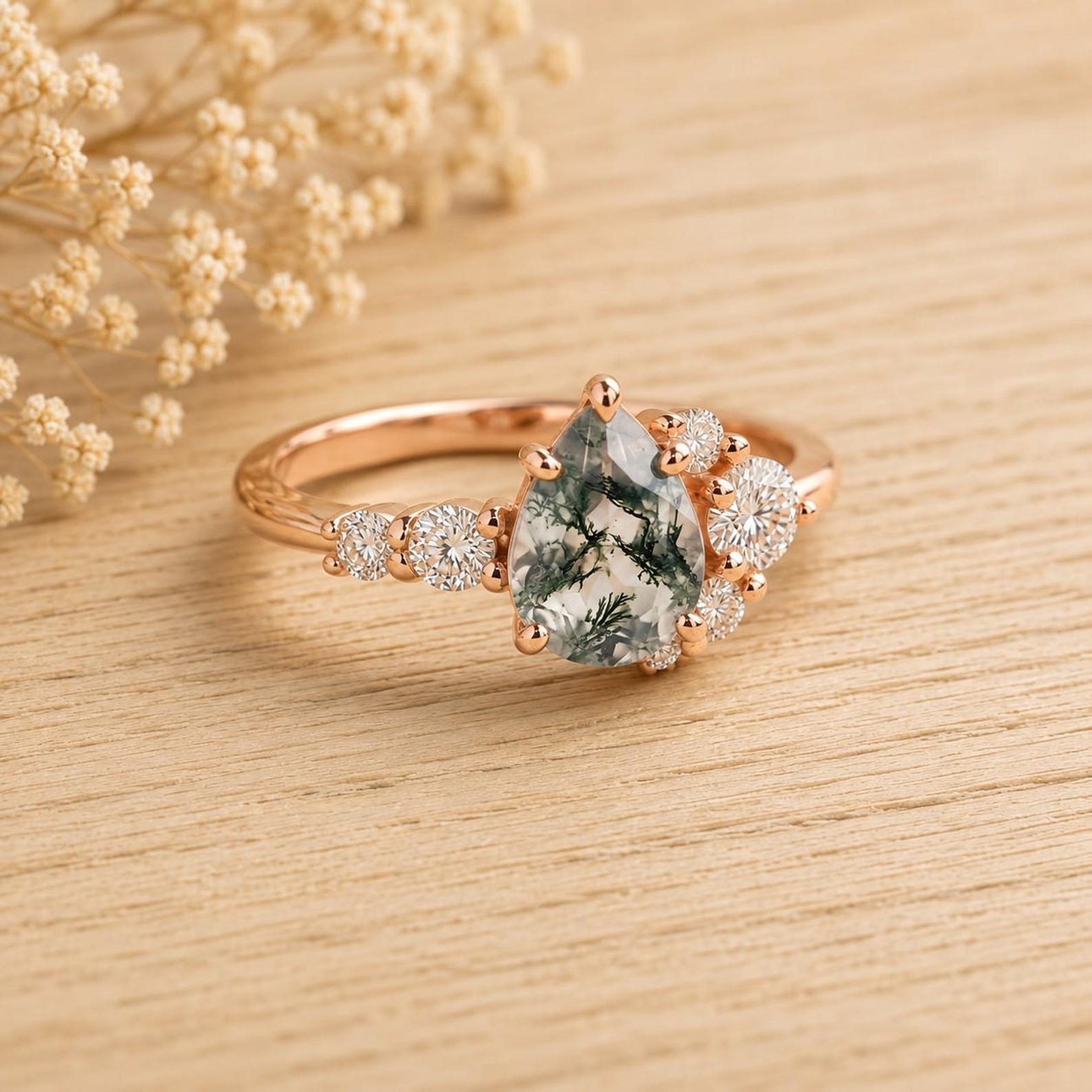 May include: A rose gold ring featuring a central pear-shaped moss agate gemstone with dark green inclusions. The stone is flanked by a cluster of small, round, clear faceted stones set along the band, creating a delicate and balanced arrangement.
