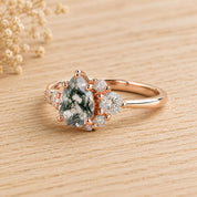 May include: A rose gold ring featuring a pear-shaped moss agate center stone with dark green dendritic inclusions. The setting is accented by a cluster of small, round, brilliant-cut white stones on one side of the pear-shaped gemstone.