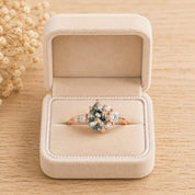 May include: A rose gold ring featuring a pear-shaped moss agate center stone with dark green inclusions, accented by a cluster of small round white stones on each side, presented inside a beige velvet jewelry box with a hinged lid.