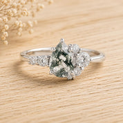May include: A silver-toned ring featuring a central pear-shaped moss agate gemstone with dark green fern-like inclusions. The stone is flanked by a cluster of small, round, clear faceted stones on both sides, set on a polished metal band.