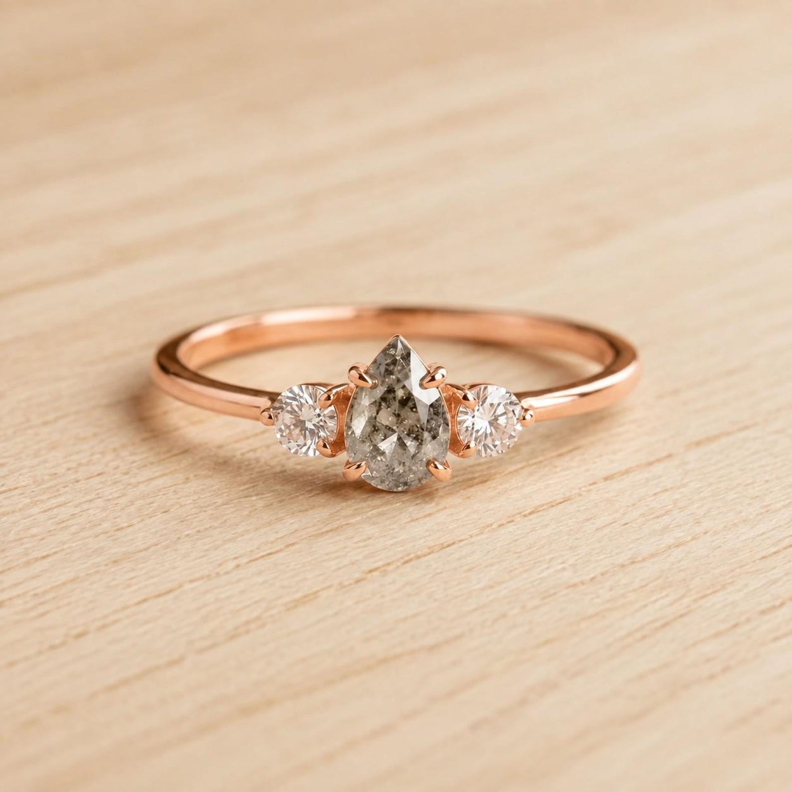 May include: A rose gold ring featuring a central pear-shaped salt and pepper diamond flanked by two smaller round brilliant-cut white diamonds. The stones are set in a delicate metal band with a polished finish and secure prong settings.