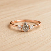 May include: A rose gold ring featuring a central pear-shaped salt and pepper diamond flanked by two smaller round brilliant-cut white diamonds. The stones are set in a delicate metal band with a polished finish and secure prong settings.