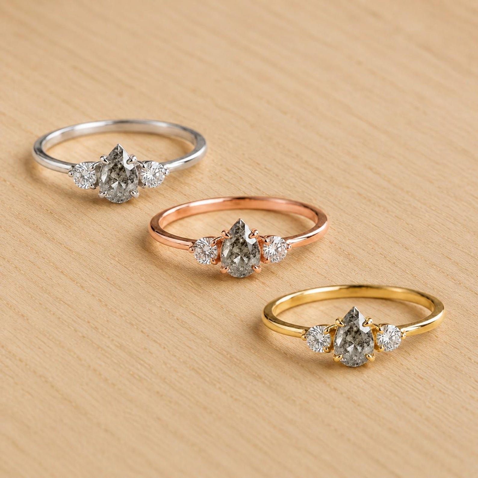 May include: Three rings featuring a central pear-shaped salt and pepper diamond flanked by two small round brilliant-cut diamonds. The rings are crafted in silver-toned, rose gold-toned, and yellow gold-toned metal bands with prong settings.